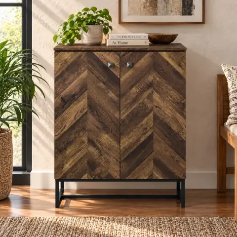 Two door wooden cabinet shown closed in a warm sunlit interior with dark chevron-patterned fronts, square knobs and a black metal base.