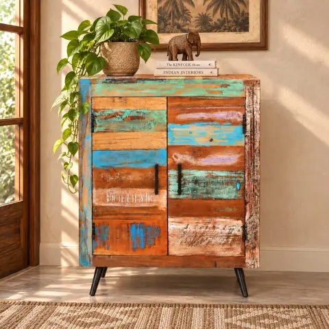 Distressed multicolor wooden storage cabinet with two closed doors, black vertical handles and slim dark legs styled in a warm interior.