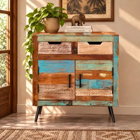 Distressed multicolor wood storage cabinet shown closed in a warm interior, featuring two upper drawers, two lower doors, dark metal handles and angled black legs.