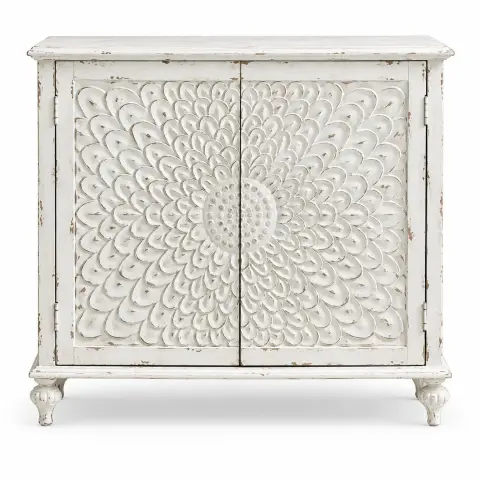 Distressed white wooden storage cabinet with two carved floral mandala doors and turned feet styled in a warm living room interior.