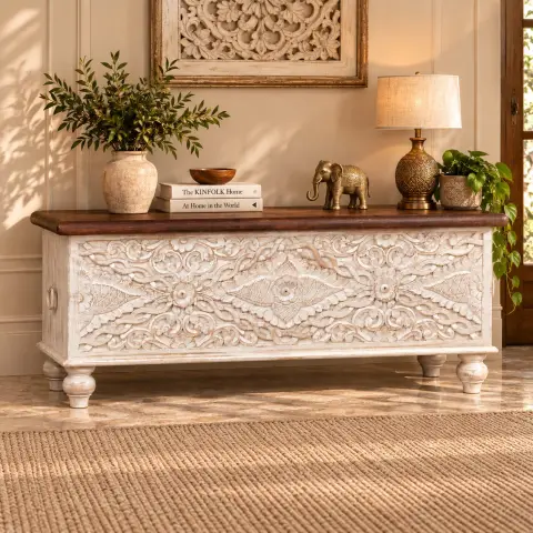 Hand-carved mango wood blanket box in distressed white finish with dark wood top, floral carved front, turned legs and side handles in a warm interior setting.