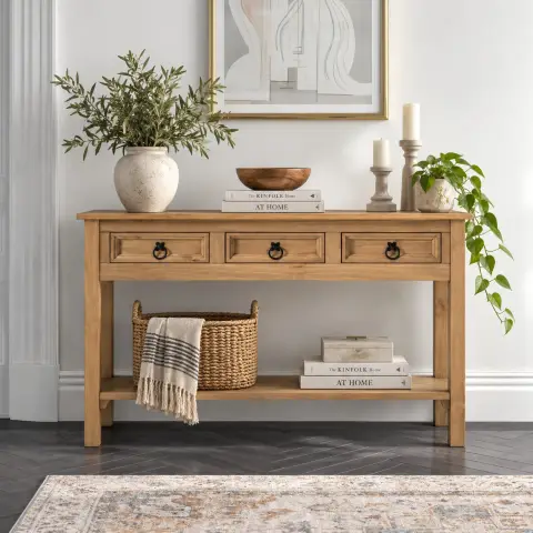 Three drawer wooden console table with dark ring pulls and a full-width lower shelf styled with plants and decorative objects in a bright interior.